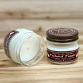 Two 4 ounce glass square mason jar candles with brown lids and labels on a wooden surface. One candle is upright, the other is tilted and open showing the candles white wax. The labels read 'Bear Family Candles, Orchard Treats with scent details: Cinnamon - Apple - Vanilla. Hand Poured Soy Candle’. The lid features a brown bear logo.