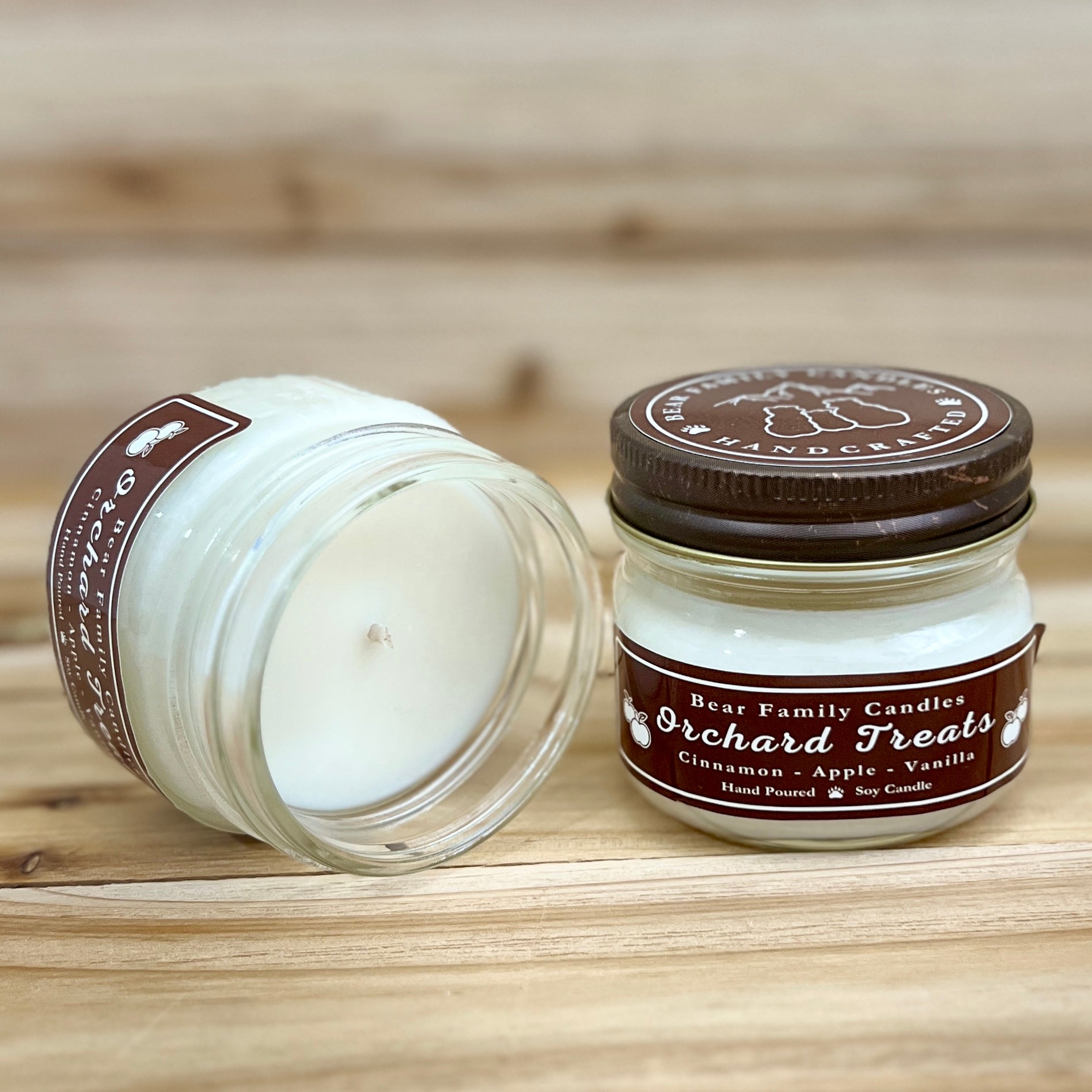 Two 4 ounce glass square mason jar candles with brown lids and labels on a wooden surface. One candle is upright, the other is tilted and open showing the candles white wax. The labels read 'Bear Family Candles, Orchard Treats with scent details: Cinnamon - Apple - Vanilla. Hand Poured Soy Candle’. The lid features a brown bear logo.