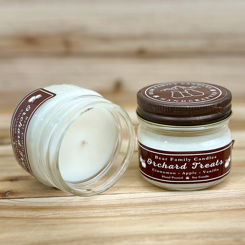 Two 4 ounce glass square mason jar candles with brown lids and labels on a wooden surface. One candle is upright, the other is tilted and open showing the candles white wax. The labels read 'Bear Family Candles, Orchard Treats with scent details: Cinnamon - Apple - Vanilla. Hand Poured Soy Candle’. The lid features a brown bear logo.