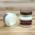 Two 8 ounce glass square mason jar candles with brown lids and labels on a wooden surface. One candle is upright, the other is tilted and open showing the candles white wax. The labels read 'Bear Family Candles, Orchard Treats with scent details: Cinnamon - Apple - Vanilla. Hand Poured Soy Candle’. The lid features a brown bear logo.