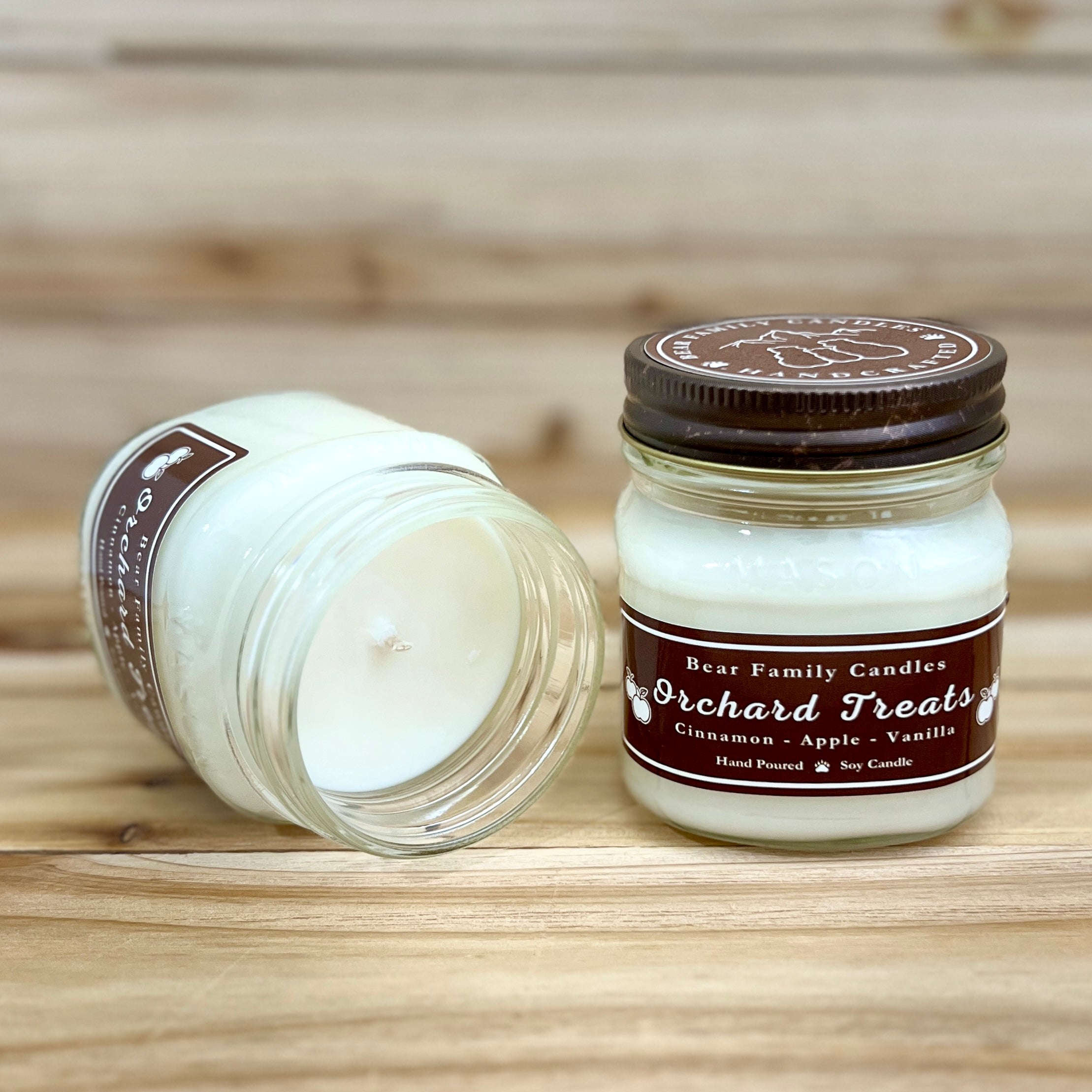 Two 8 ounce glass square mason jar candles with brown lids and labels on a wooden surface. One candle is upright, the other is tilted and open showing the candles white wax. The labels read 'Bear Family Candles, Orchard Treats with scent details: Cinnamon - Apple - Vanilla. Hand Poured Soy Candle’. The lid features a brown bear logo.