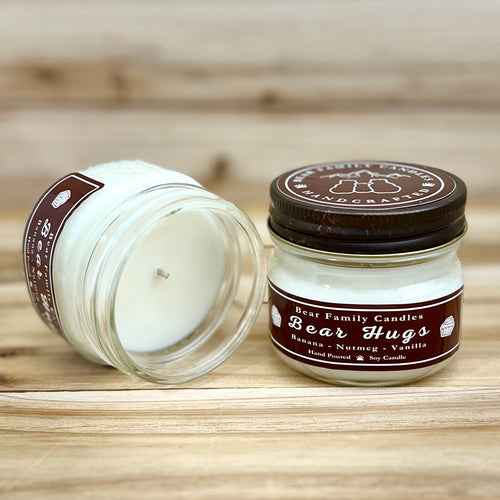 Two 4 ounce glass square mason jar candles with brown lids and labels on a wooden surface. One candle is upright, the other is tilted and open showing the candles white wax The labels read "Bear Family Candles, Bear Hugs" with scent details: Banana, Nutmeg, Vanilla. Hand poured soy candle. The lid features a brown bear logo.
