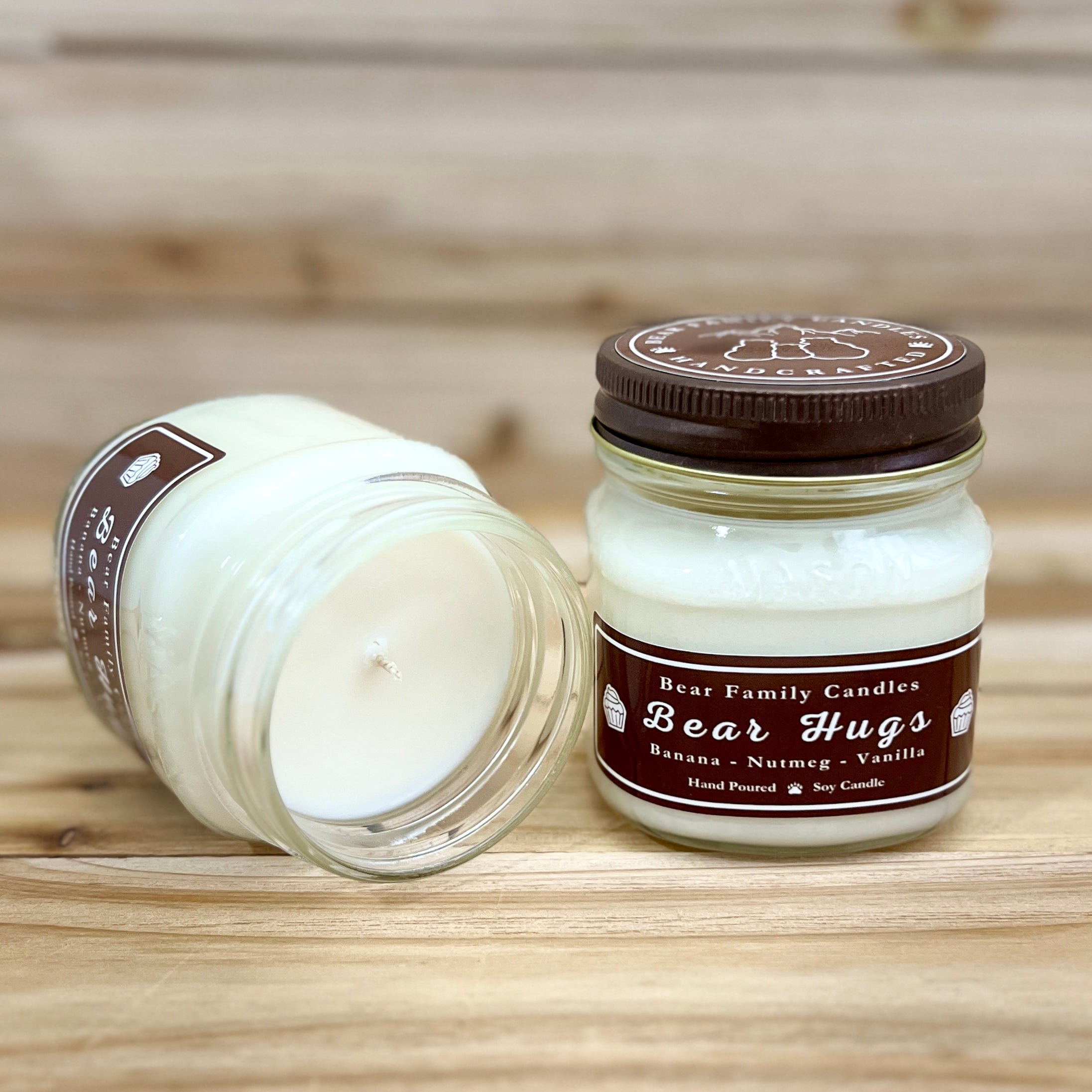 Two 8 ounce glass square mason jar candles with brown lids and labels on a wooden surface. One candle is upright, the other is tilted and open showing the candles white wax The labels read "Bear Family Candles, Bear Hugs" with scent details: Banana, Nutmeg, Vanilla. Hand poured soy candle. The lid features a brown bear logo.