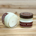 Two 4 ounce glass square mason jar candles with brown lids and labels on a wooden surface. One candle is upright, the other is tilted and open showing the candles white wax. The labels read "Bear Family Candles, Little Bear" with scent details: cool mint and creamy vanilla. Hand poured soy candle. The lid features a brown bear logo.