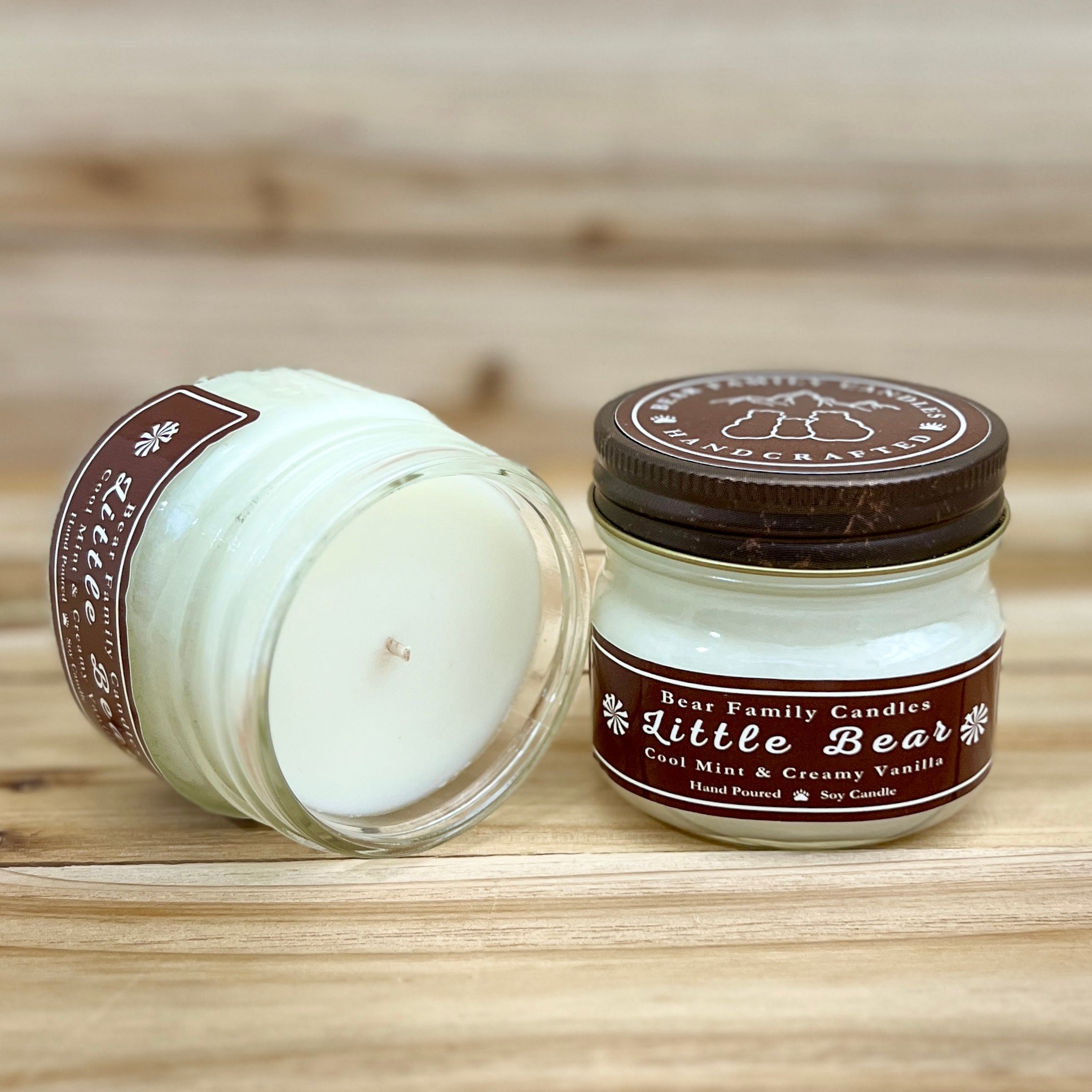 Two 4 ounce glass square mason jar candles with brown lids and labels on a wooden surface. One candle is upright, the other is tilted and open showing the candles white wax. The labels read "Bear Family Candles, Little Bear" with scent details: cool mint and creamy vanilla. Hand poured soy candle. The lid features a brown bear logo.