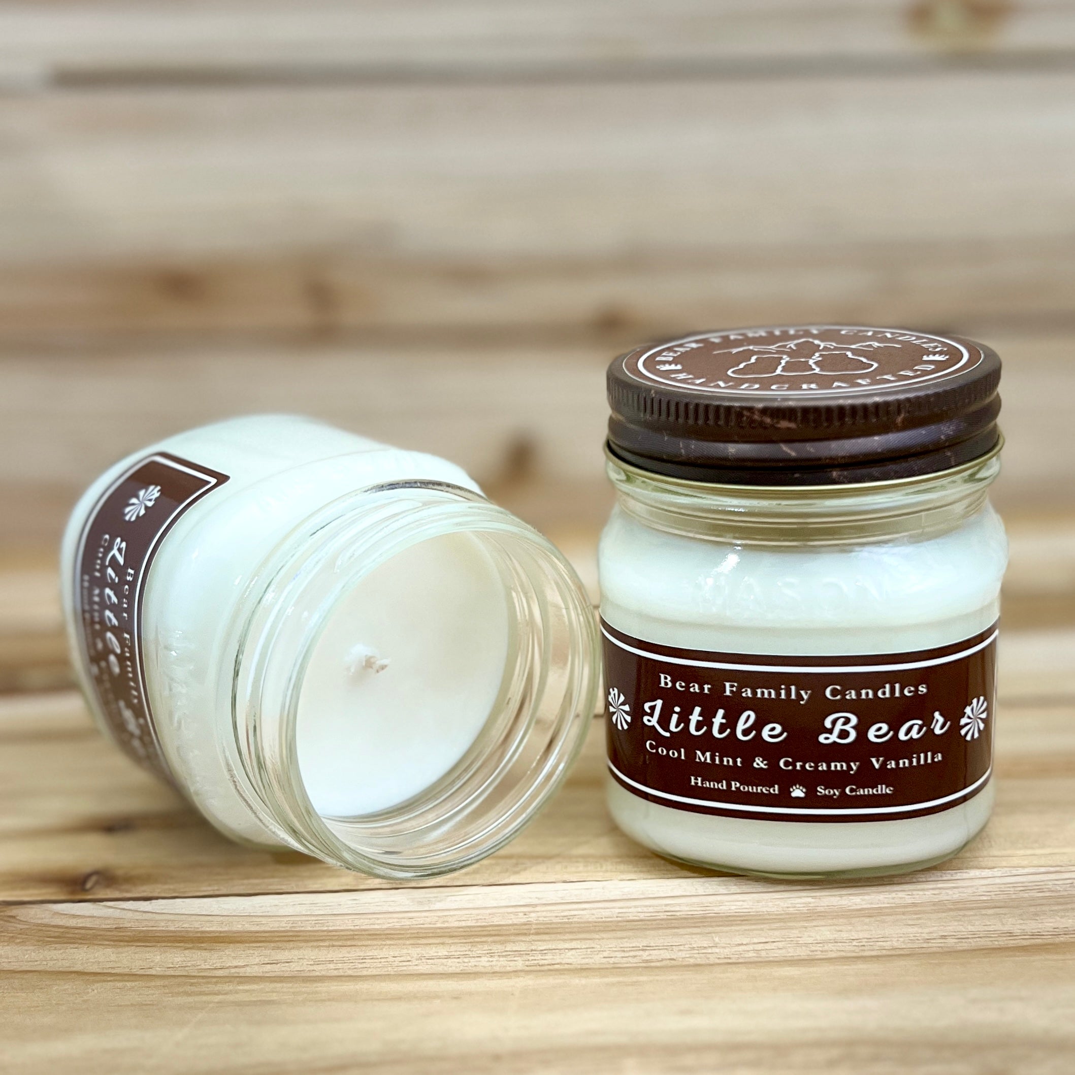 Two 8 ounce glass square mason jar candles with brown lids and labels on a wooden surface. One candle is upright, the other is tilted and open showing the candles white wax. The labels read "Bear Family Candles, Little Bear" with scent details: cool mint and creamy vanilla. Hand poured soy candle. The lid features a brown bear logo.