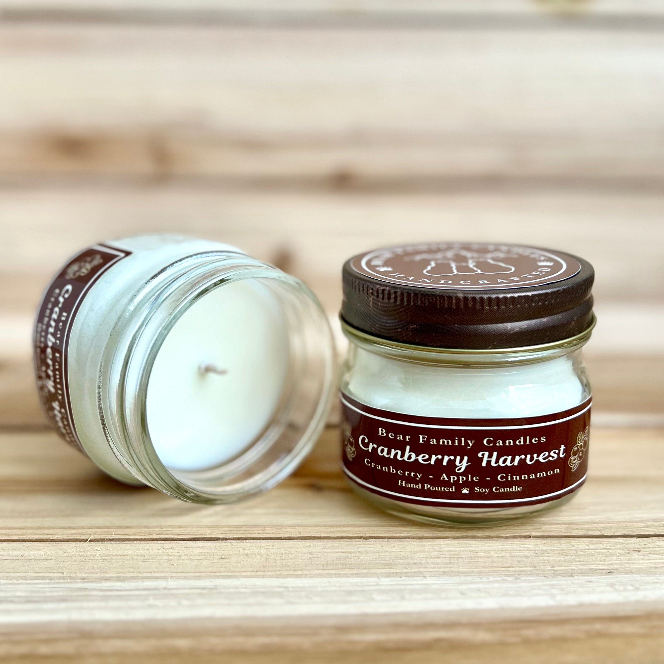 Two 4 ounce glass square mason jar candles with brown lids and labels on a wooden surface. One candle is upright, the other is tilted and open showing the candles white wax. The labels read "Bear Family Candles, Cranberry Harvest" with scent details: cranberry apple cinnamon. Hand poured soy candle. The lid features a brown bear logo.