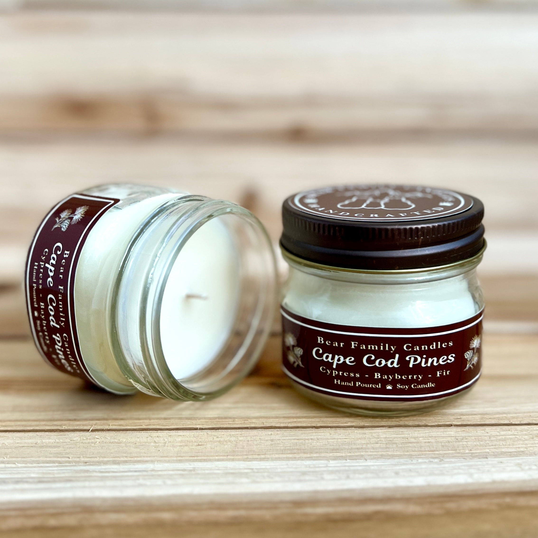 Two 4 ounce glass square mason jar candles with brown lids and labels on a wooden surface. One candle is upright, the other is tilted and open showing the candles white wax. The labels read "Bear Family Candles, Cape Cod Pines" with scent details: cypress bayberry fir. Hand poured soy candle. The lid features a brown bear logo.