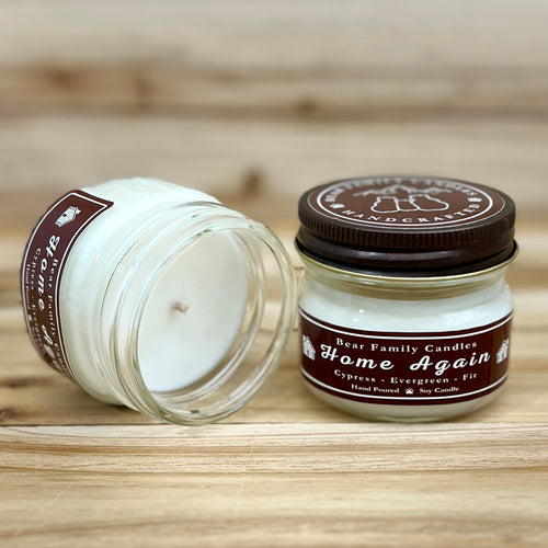 Two 4 ounce glass square mason jar candles with brown lids and labels on a wooden surface. One candle is upright, the other is tilted and open showing the candles white wax. The labels read "Bear Family Candles, Home Again" with scent details: Cypress, Evergreen, and Fir. Hand poured soy candle. The lid features a brown bear logo.