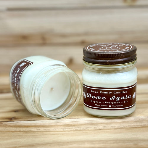 Two 8 ounce glass square mason jar candles with brown lids and labels on a wooden surface. One candle is upright, the other is tilted and open showing the candles white wax. The labels read "Bear Family Candles, Home Again" with scent details: Cypress, Evergreen, and Fir. Hand poured soy candle. The lid features a brown bear logo.