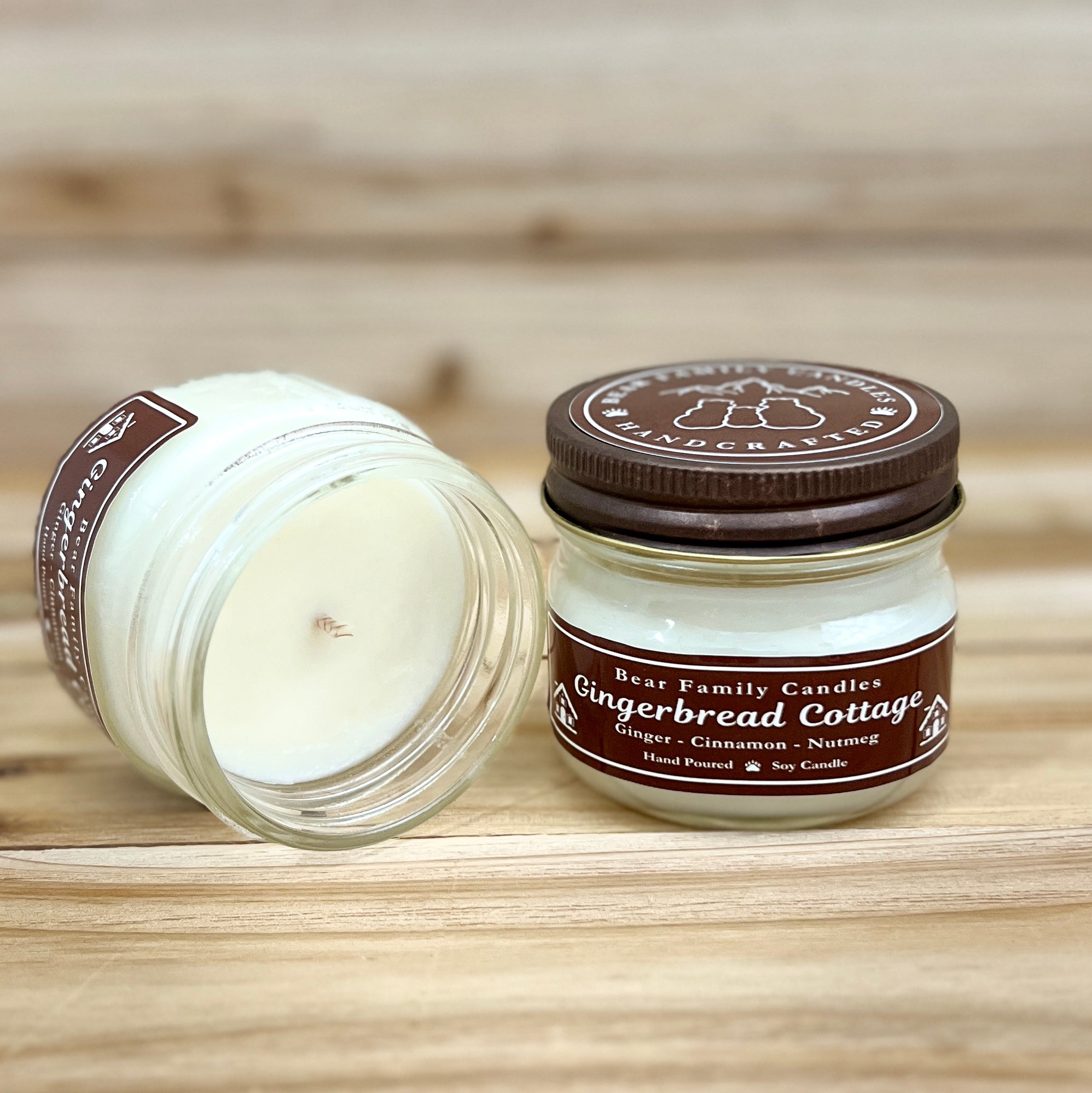 Two 4 ounce glass square mason jar candles with brown lids and labels on a wooden surface. One candle is upright, the other is tilted and open showing the candles white wax. The labels read "Bear Family Candles, Gingerbread Cottage" with scent details: Ginger cinnamon nutmeg. Hand poured soy candle. The lid features a brown bear logo.