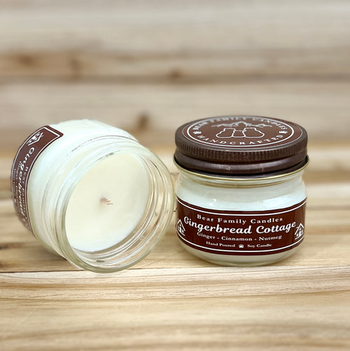 Two 4 ounce glass square mason jar candles with brown lids and labels on a wooden surface. One candle is upright, the other is tilted and open showing the candles white wax. The labels read "Bear Family Candles, Gingerbread Cottage" with scent details: Ginger cinnamon nutmeg. Hand poured soy candle. The lid features a brown bear logo.