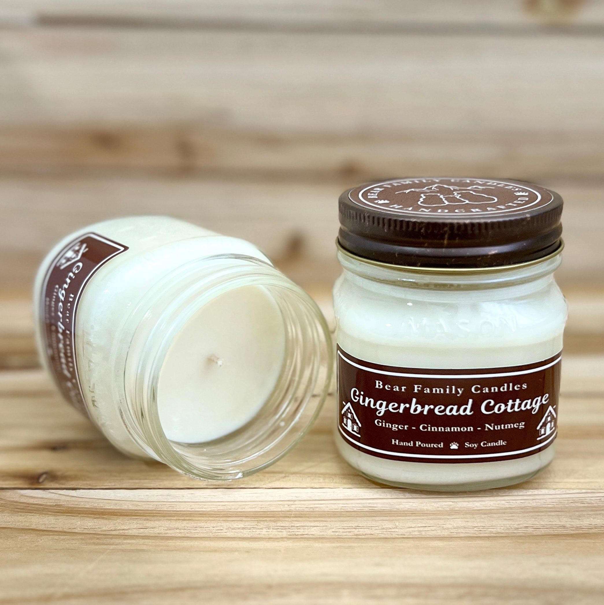 Two 8 ounce glass square mason jar candles with brown lids and labels on a wooden surface. One candle is upright, the other is tilted and open showing the candles white wax. The labels read "Bear Family Candles, Gingerbread Cottage" with scent details: Ginger cinnamon nutmeg. Hand poured soy candle. The lid features a brown bear logo.