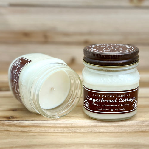 Two 8 ounce glass square mason jar candles with brown lids and labels on a wooden surface. One candle is upright, the other is tilted and open showing the candles white wax. The labels read "Bear Family Candles, Gingerbread Cottage" with scent details: Ginger cinnamon nutmeg. Hand poured soy candle. The lid features a brown bear logo.