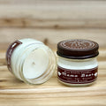 Two 4 ounce glass square mason jar candles with brown lids and labels on a wooden surface. One candle is upright, the other is tilted and open showing the candles white wax. The labels read "Bear Family Candles, Mama Bear" with scent details: lavender sage rosemary. Hand poured soy candle. The lid features a brown bear logo.