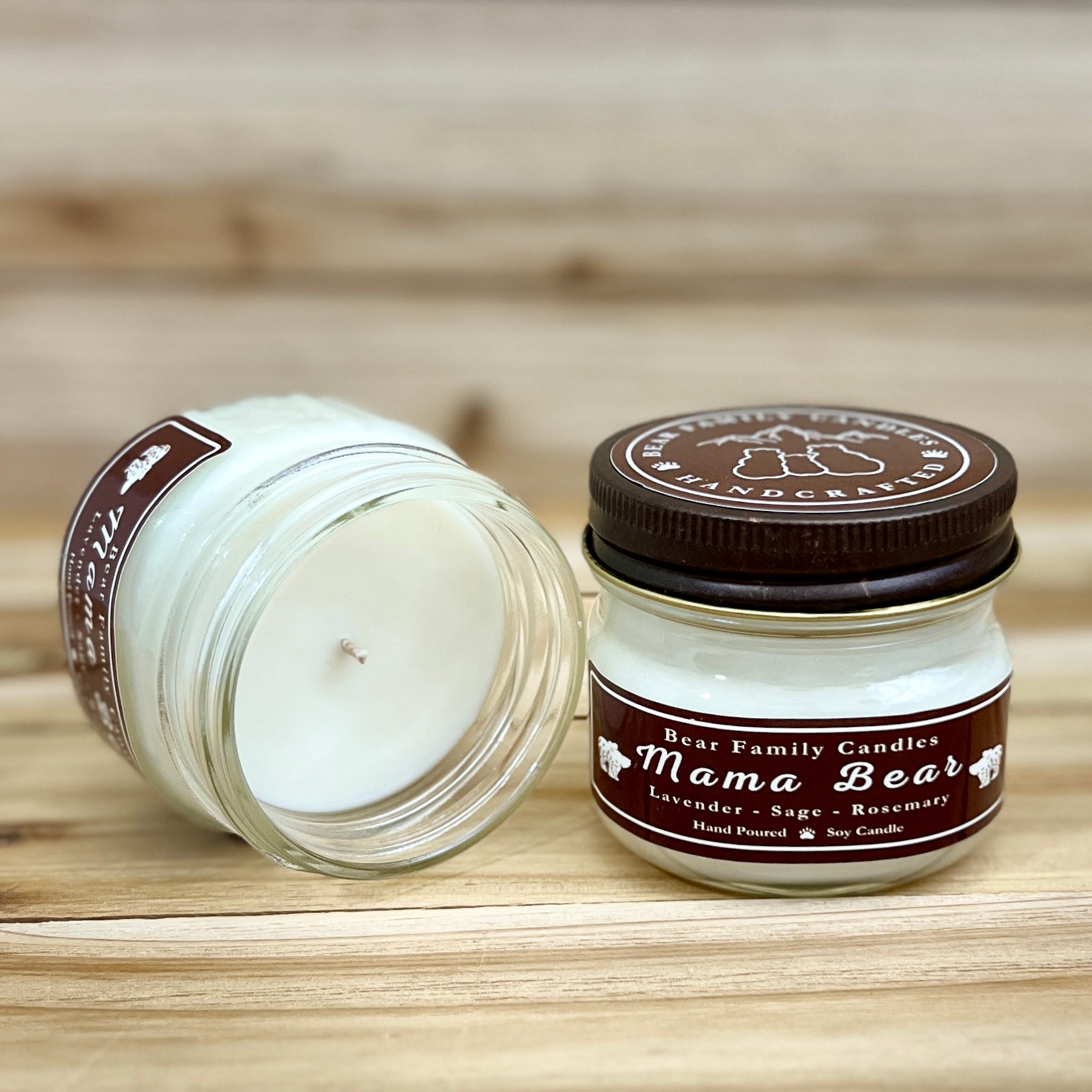 Two 4 ounce glass square mason jar candles with brown lids and labels on a wooden surface. One candle is upright, the other is tilted and open showing the candles white wax. The labels read "Bear Family Candles, Mama Bear" with scent details: lavender sage rosemary. Hand poured soy candle. The lid features a brown bear logo.