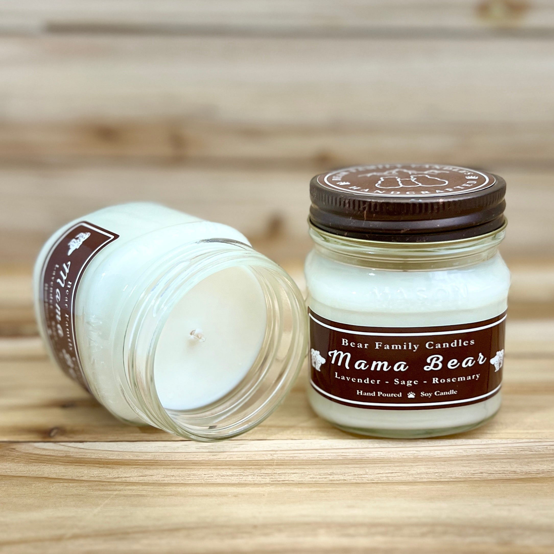 Two 8 ounce glass square mason jar candles with brown lids and labels on a wooden surface. One candle is upright, the other is tilted and open showing the candles white wax. The labels read "Bear Family Candles, Mama Bear" with scent details: lavender sage rosemary. Hand poured soy candle. The lid features a brown bear logo.