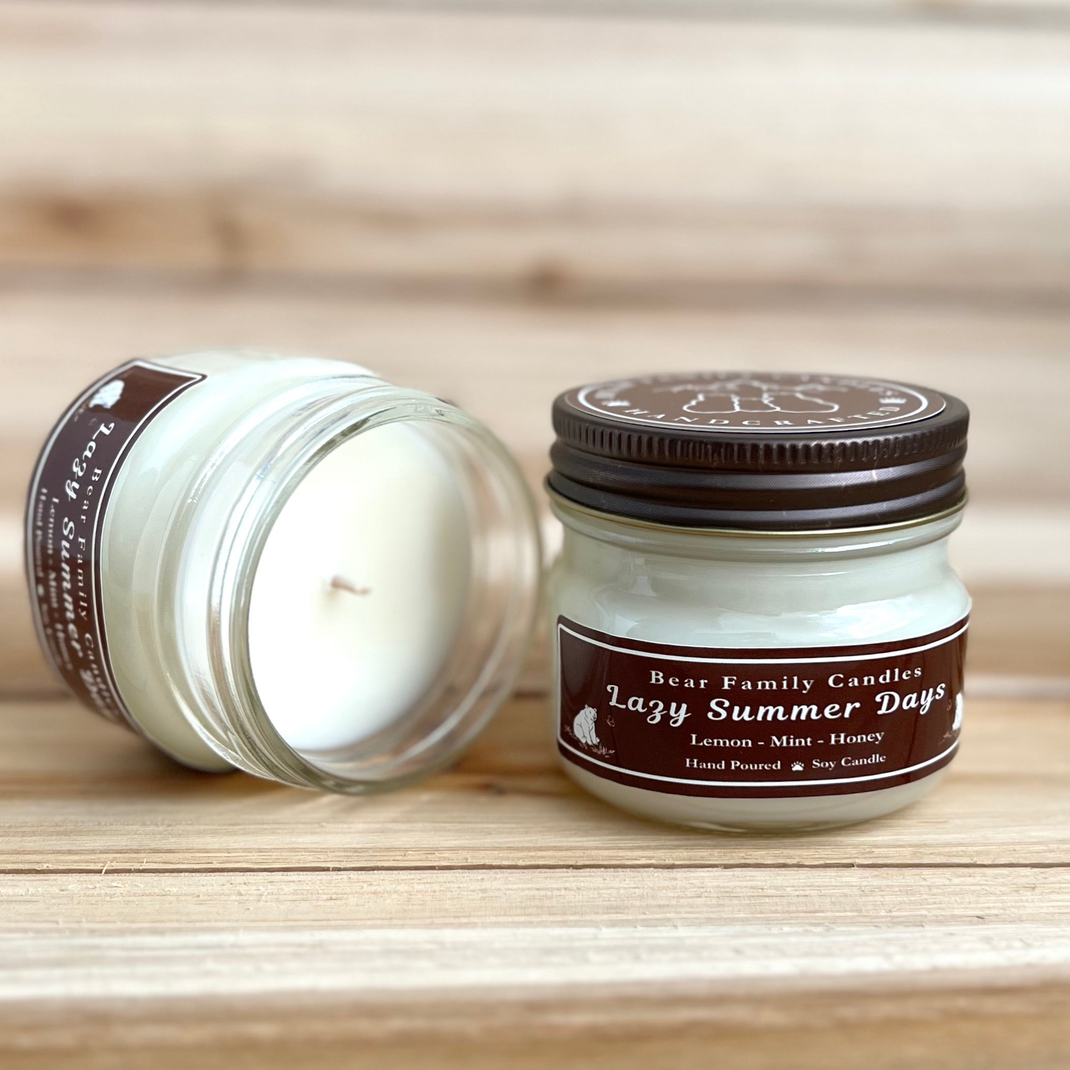 Two 4 ounce glass square mason jar candles with brown lids and labels on a wooden surface. One candle is upright, the other is tilted and open showing the candles white wax. The labels read "Bear Family Candles, Lazy summer Days" with scent details: lemon mint honey. Hand poured soy candle. The lid features a brown bear logo.