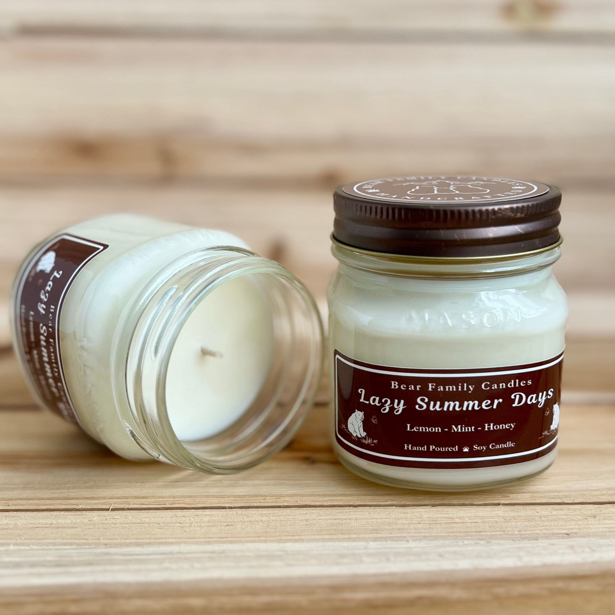 Two 8 ounce glass square mason jar candles with brown lids and labels on a wooden surface. One candle is upright, the other is tilted and open showing the candles white wax. The labels read "Bear Family Candles, Lazy Summer Days" with scent details: lemon mint honey. Hand poured soy candle. The lid features a brown bear logo.