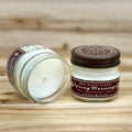 Two 4 ounce glass square mason jar candles with brown lids and labels on a wooden surface. One candle is upright, the other is tilted and open showing the candles white wax. The labels read "Bear Family Candles, Spring Mornings" with scent details: lilac honeysuckle amyris. Hand poured soy candle. The lid features a brown bear logo.