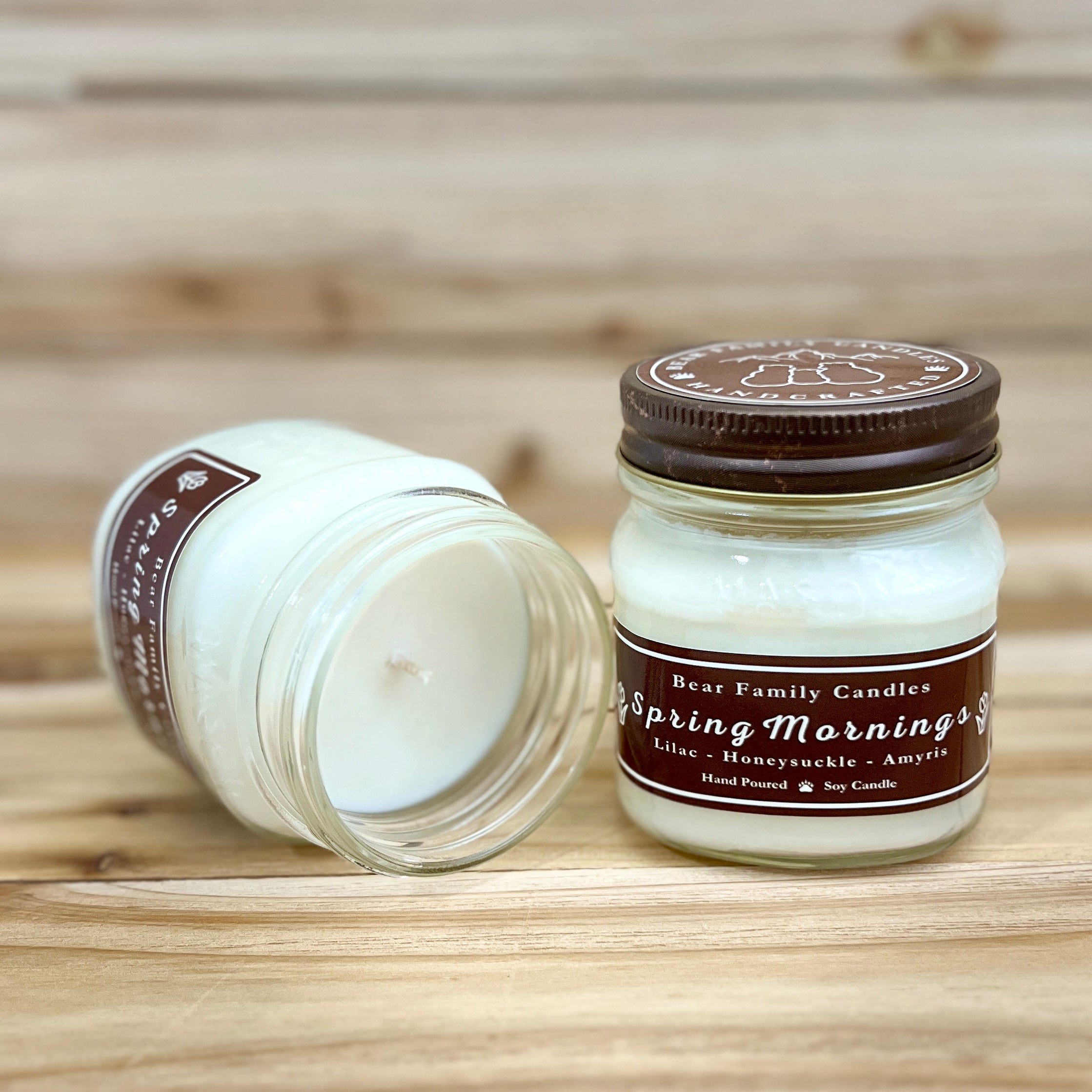 Two 8 ounce glass square mason jar candles with brown lids and labels on a wooden surface. One candle is upright, the other is tilted and open showing the candles white wax. The labels read "Bear Family Candles, Spring Mornings" with scent details: lilac honeysuckle amyris. Hand poured soy candle. The lid features a brown bear logo.