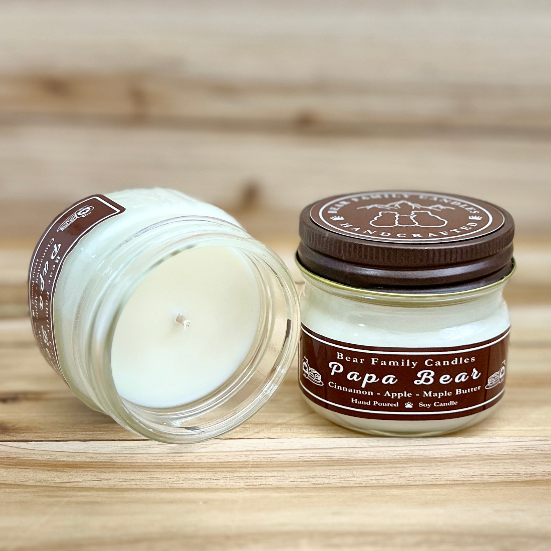 Two 4 ounce glass square mason jar candles with brown lids and labels on a wooden surface. One candle is upright, the other is tilted and open showing the candles white wax. The labels read "Bear Family Candles, Papa Bear" with scent details: Cinnamon Apple Maple Butter. Hand poured soy candle. The lid features a brown bear logo.