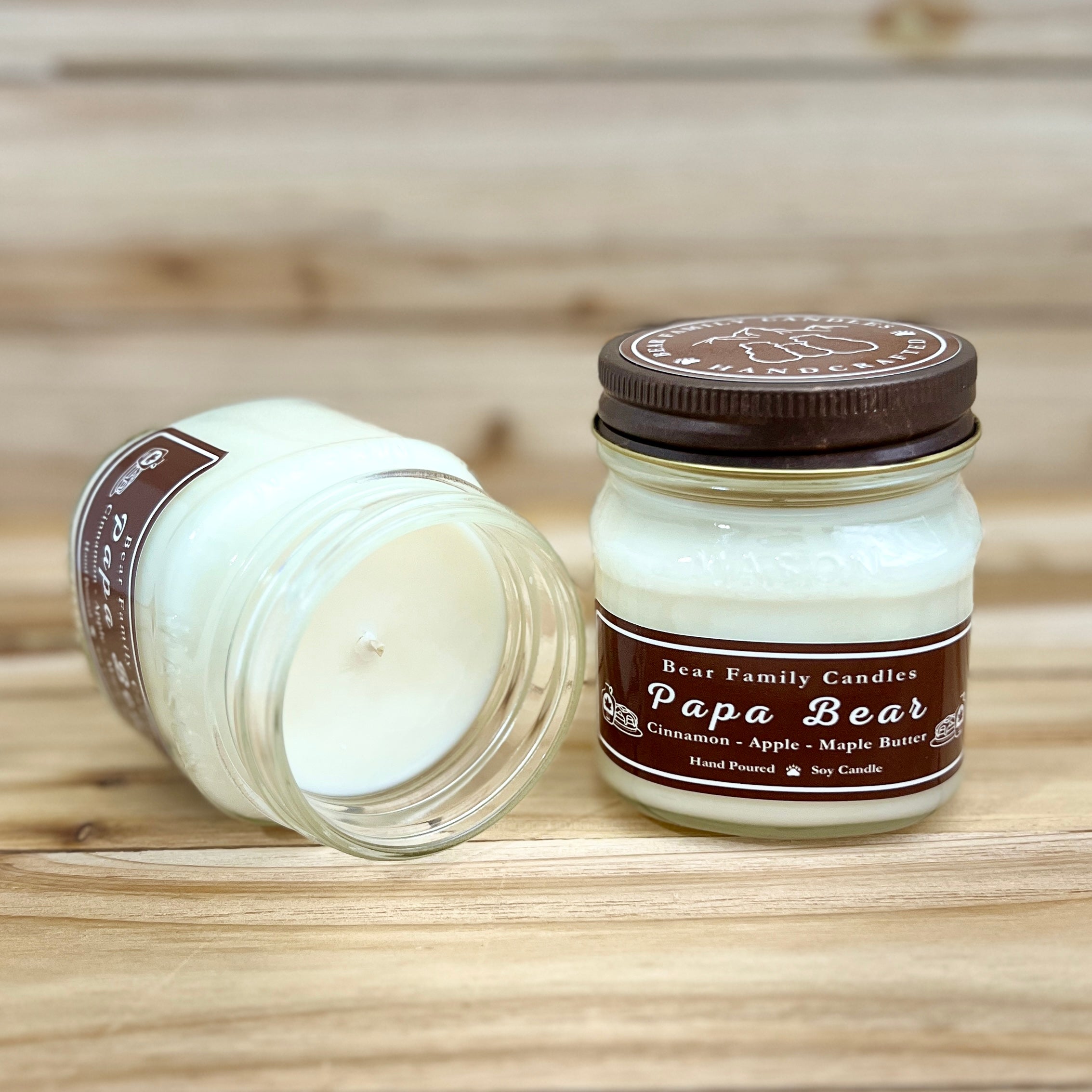 Two 8 ounce glass square mason jar candles with brown lids and labels on a wooden surface. One candle is upright, the other is tilted and open showing the candles white wax. The labels read "Bear Family Candles, Papa Bear" with scent details: maple apple cinnamon. Hand poured soy candle. The lid features a brown bear logo.