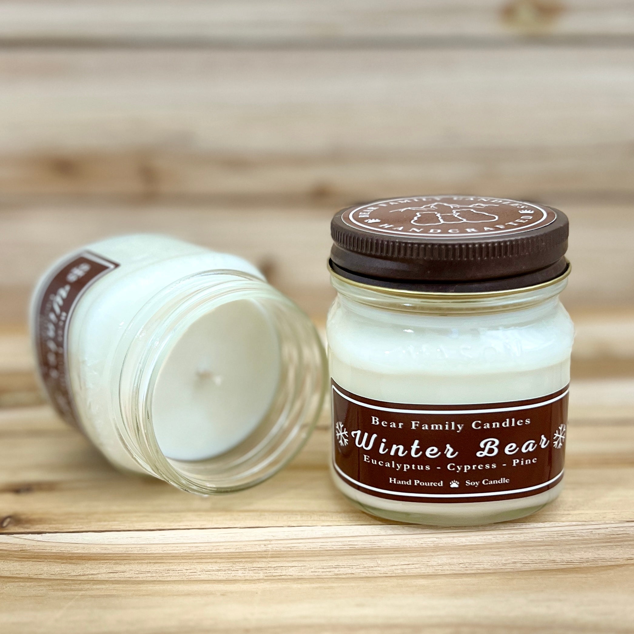 Two 8 ounce glass square mason jar candles with brown lids and labels on a wooden surface. One candle is upright, the other is tilted and open showing the candles white wax. The labels read "Bear Family Candles, Winter Bear with scent details: Eucalyptus Cypress Pine. Hand Poured Soy Candle". The lid features a brown bear logo.