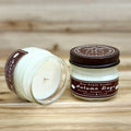 Two 4 ounce glass square mason jar candles with brown lids and labels on a wooden surface. One candle is upright, the other is tilted and open showing the candles white wax. The labels read "Bear Family Candles, Autumn Days" with scent details: nutmeg pumpkin vanilla. Hand poured soy candle. The lid features a brown bear logo.