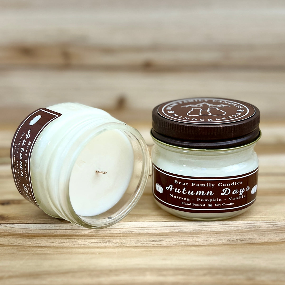 Two 4 ounce glass square mason jar candles with brown lids and labels on a wooden surface. One candle is upright, the other is tilted and open showing the candles white wax. The labels read "Bear Family Candles, Autumn Days" with scent details: nutmeg pumpkin vanilla. Hand poured soy candle. The lid features a brown bear logo.