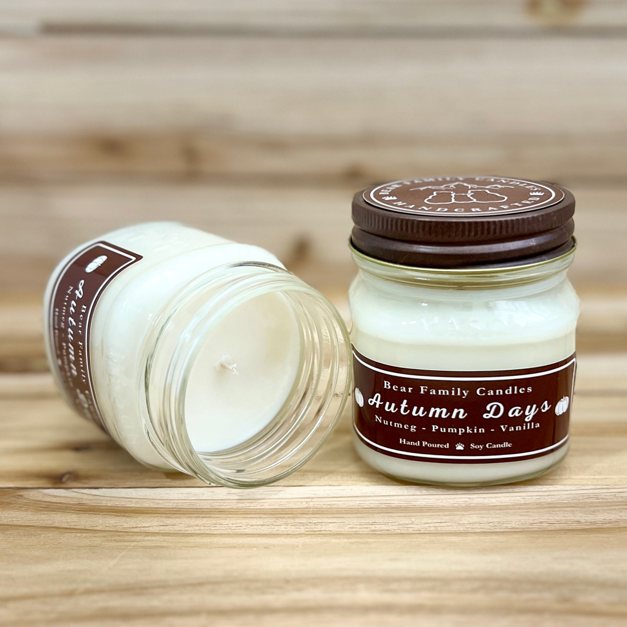 Two 8 ounce glass square mason jar candles with brown lids and labels on a wooden surface. One candle is upright, the other is tilted and open showing the candles white wax. The labels read "Bear Family Candles, Autumn Days" with scent details: nutmeg pumpkin vanilla. Hand poured soy candle. The lid features a brown bear logo.